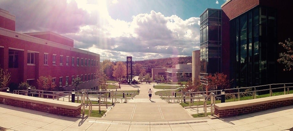 A university quad on a sunny day, brick and glass buildings either side of a paved walk with a clock tower at the far end
