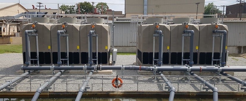 Two banks of cooling towers outdoors, joined by galvanised pipework above a water basin, with an orange life ring on the rail