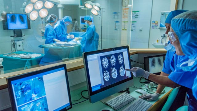 An operating theatre seen from an adjoining control room, staff in blue scrubs reviewing CT brain scans and chest X-rays on monitors while surgery goes on behind the glass