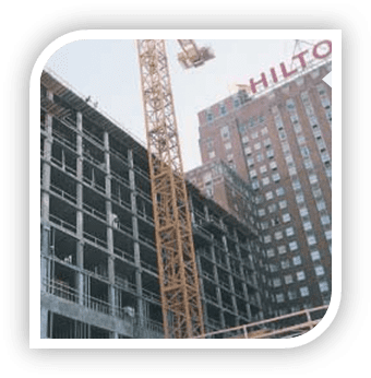 A concrete-frame building under construction with a tower crane alongside, the Hilton sign visible on the building behind