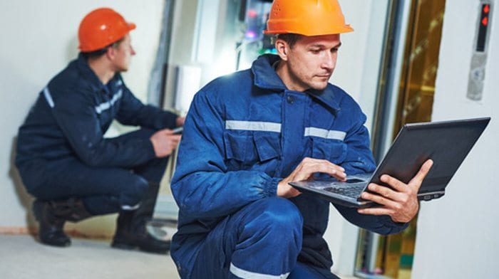 Two technicians in overalls and hard hats working in a plant room, the nearer one crouched with a laptop