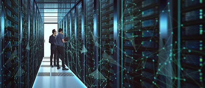Two people with a laptop standing in the aisle of a dark data hall, a network graphic overlaid across the image