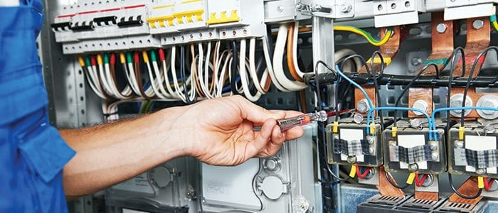 A hand using a screwdriver on terminals in an electrical distribution cabinet, breakers and coloured wiring around it
