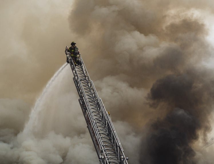 A firefighter at the top of an extended aerial ladder directing a jet of water into heavy smoke