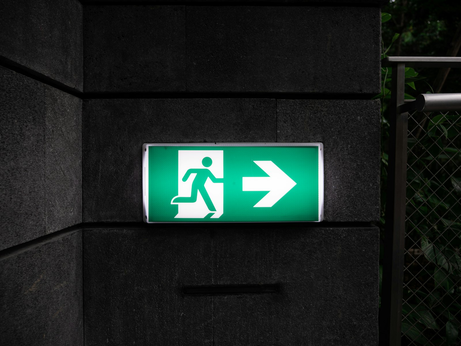 An illuminated green emergency exit sign mounted on a dark stone wall, showing a running figure and a directional arrow