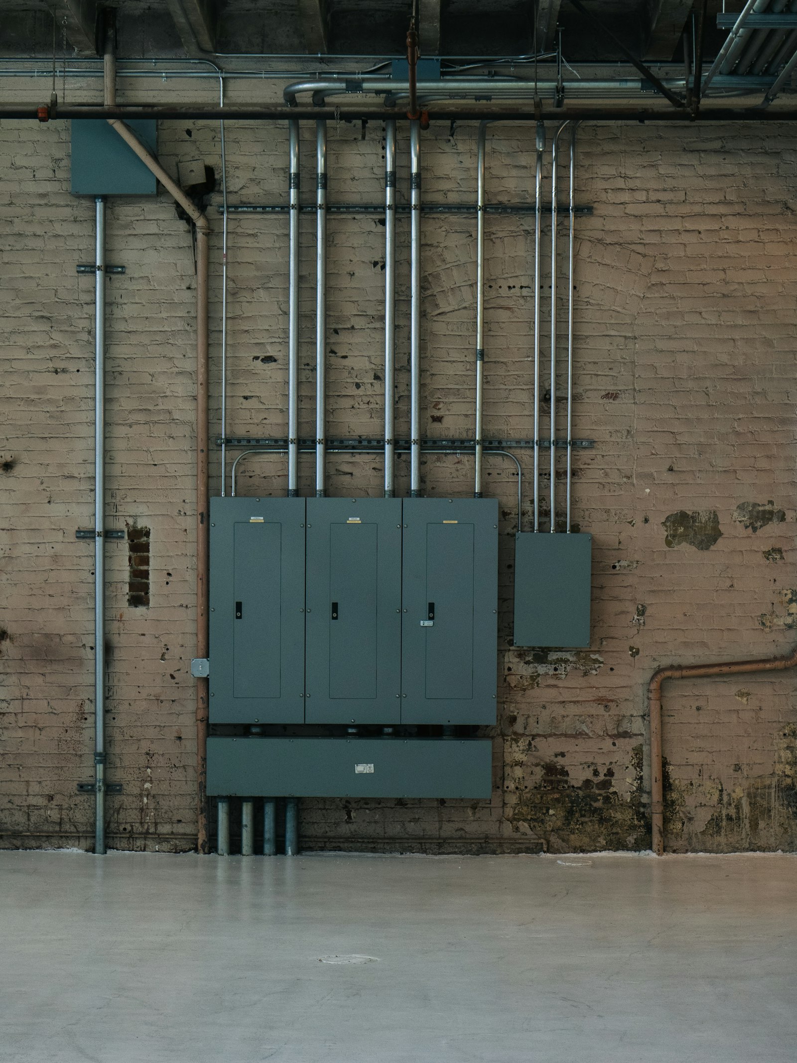 Three grey panelboards ganged together on a painted brick wall, a bank of steel conduit risers running up from them to the ceiling of an empty industrial room