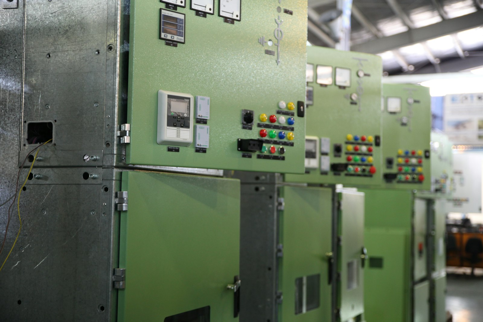 A line of green low-voltage switchgear cubicles in an industrial hall, their doors carrying analogue meters, protection relays and rows of coloured indicator lamps