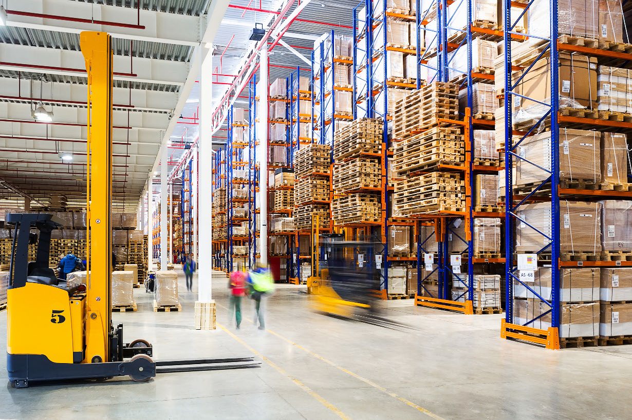 A warehouse aisle of tall pallet racking, a parked yellow reach truck in the foreground and a forklift blurred with motion as it passes