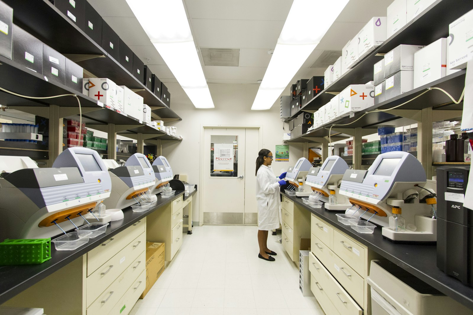 A laboratory lined on both sides with benchtop sequencing instruments, a researcher in a white coat loading a sample into one of them