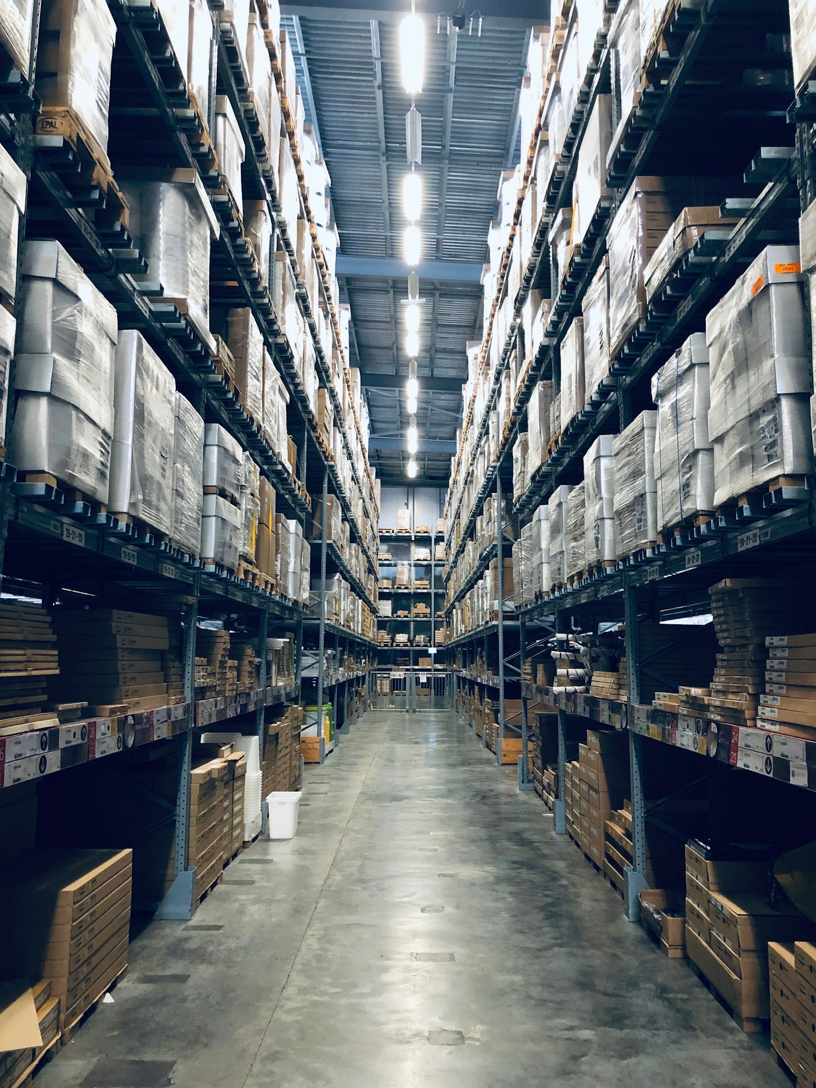 A warehouse aisle running between pallet racking stacked to the roof with shrink-wrapped goods, lit by a line of overhead fittings