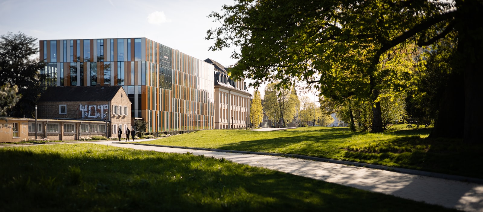 A university building with a coloured glass facade set behind mature trees and open lawn, with students walking a path in front