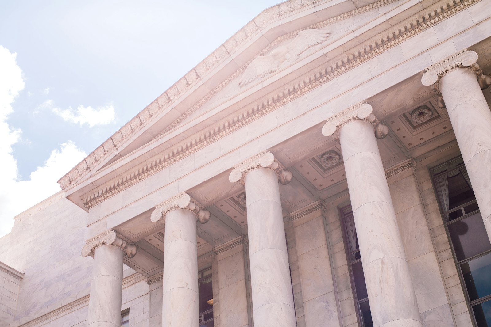 The marble portico of a neoclassical government building, looking up past Ionic columns to a carved eagle in the pediment