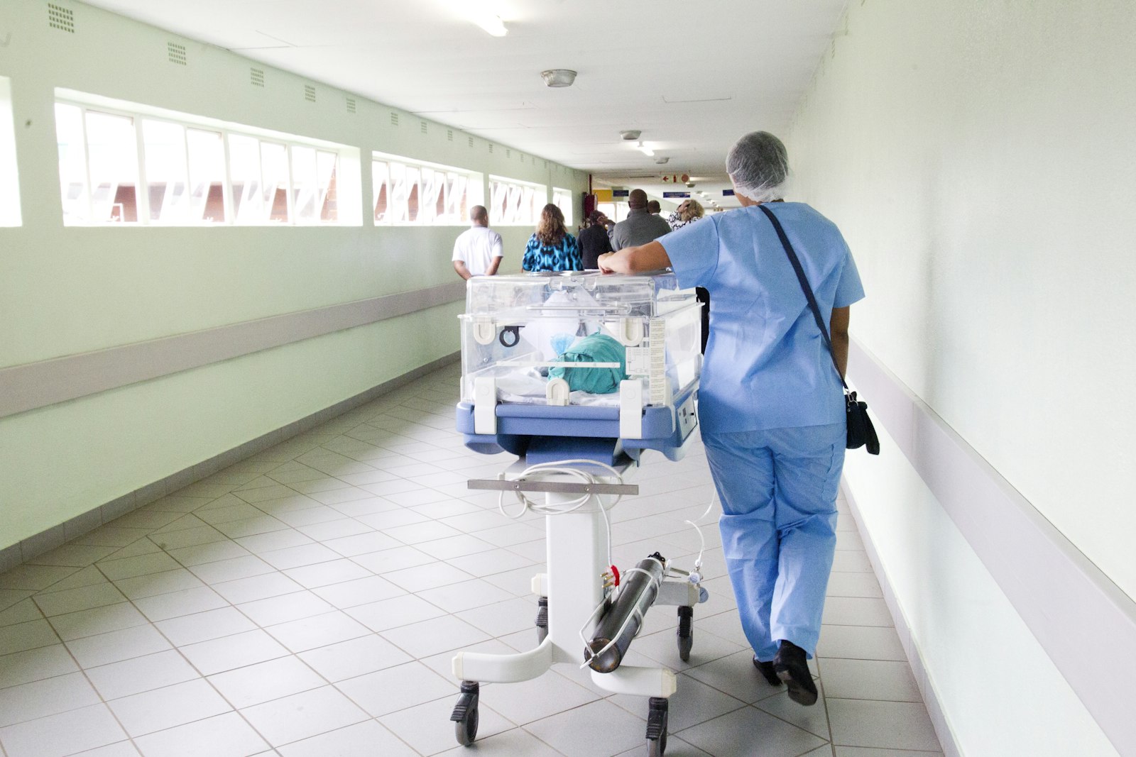 A nurse in blue scrubs wheeling an infant incubator down a bright hospital corridor, with an oxygen cylinder strapped beneath it
