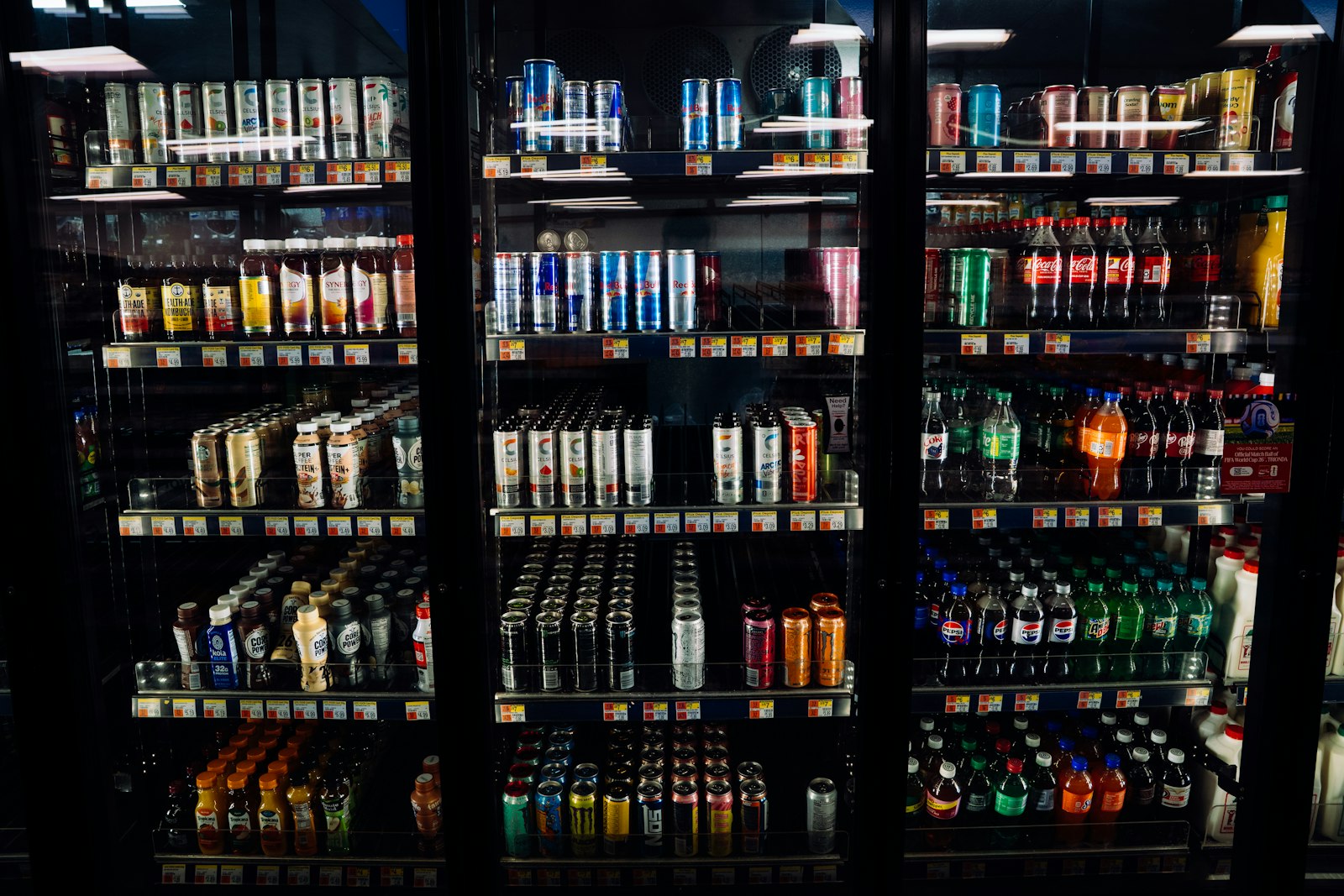 Three glass-fronted refrigerated cases in a shop, stocked from top to bottom with chilled drinks and price labels on every shelf
