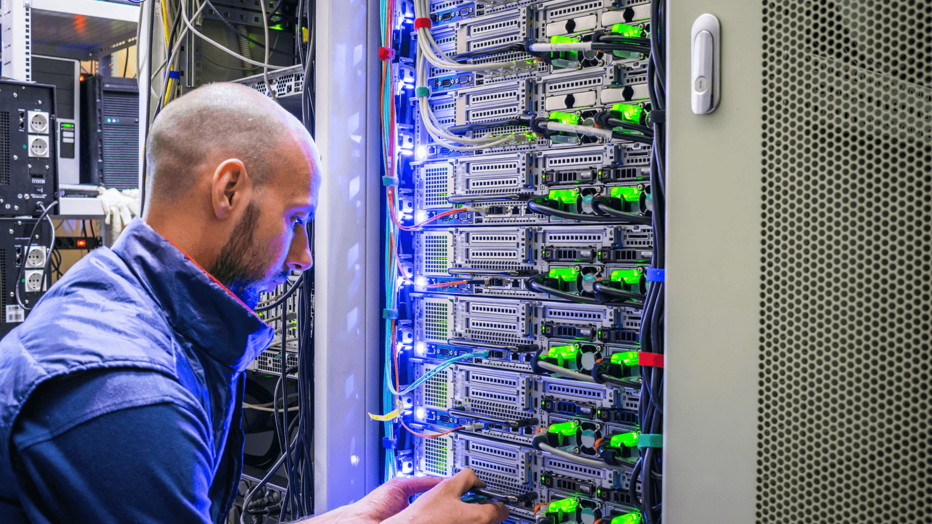 A man working at the back of a server rack, one hand among bundled patch leads and green-lit network ports
