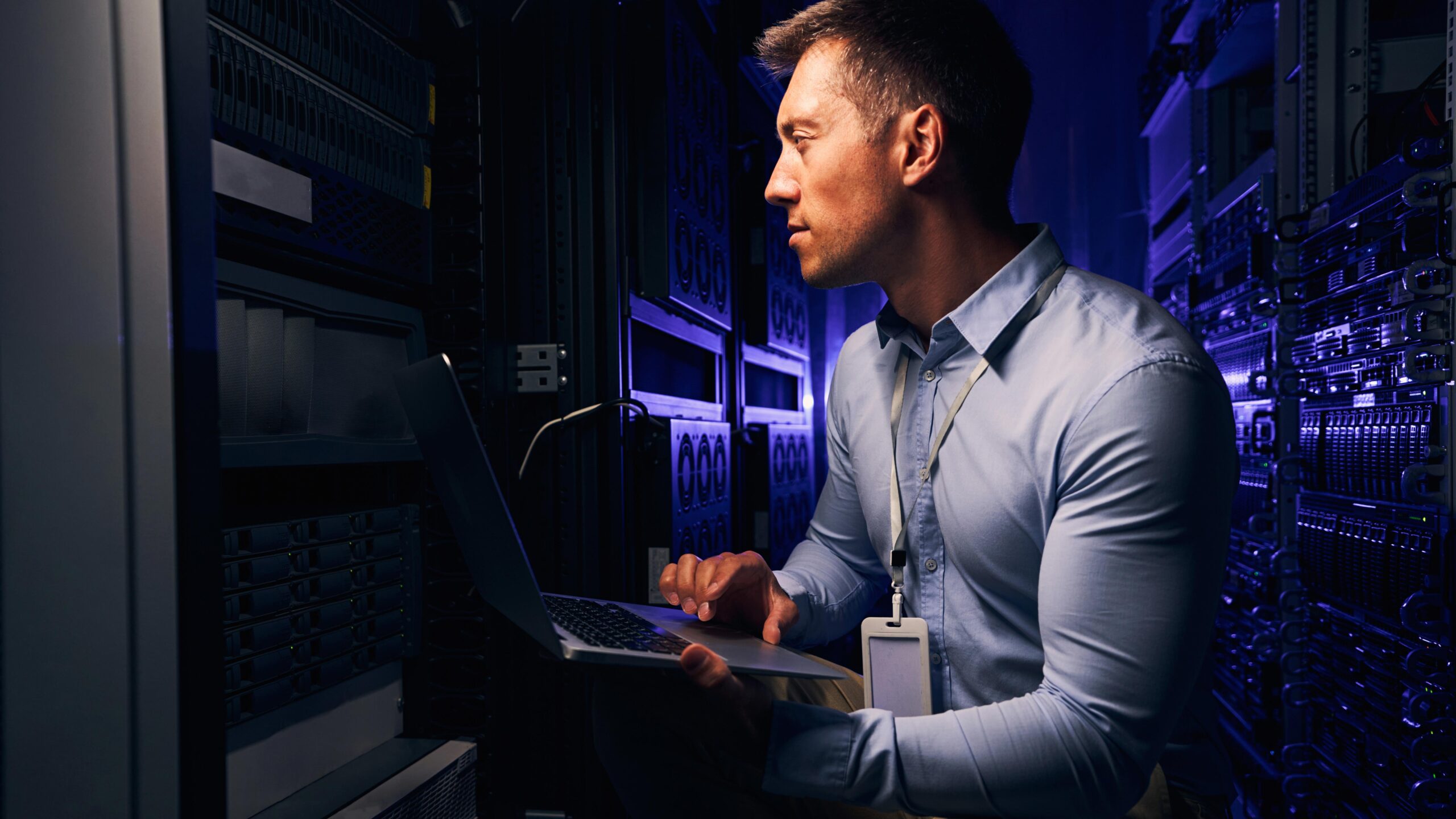 A man wearing a lanyard holds an open laptop beside a rack in a blue-lit data hall
