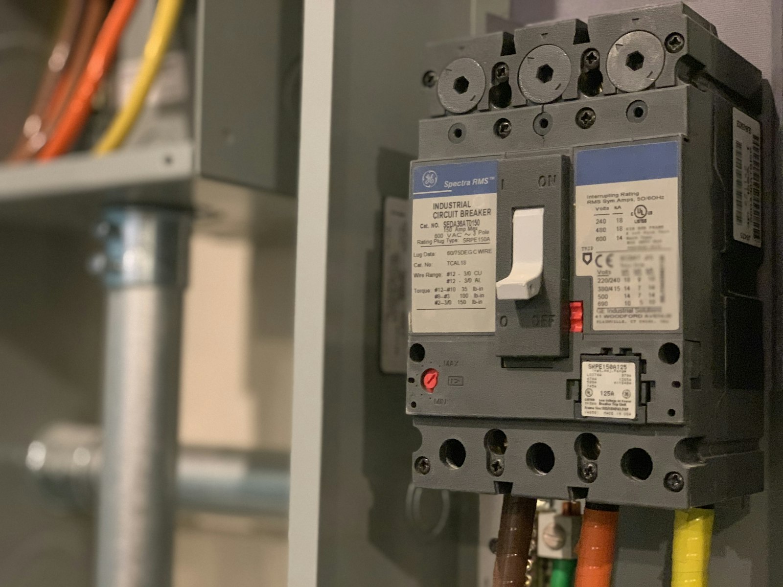 A close-up of a three-pole moulded-case industrial circuit breaker in a panel, its rating plate and interrupting-rating label legible, brown, orange and yellow phase conductors landed on the lugs below