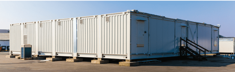A row of white containerised equipment modules on concrete plinths outdoors, a steel stair leading to a personnel door at one end