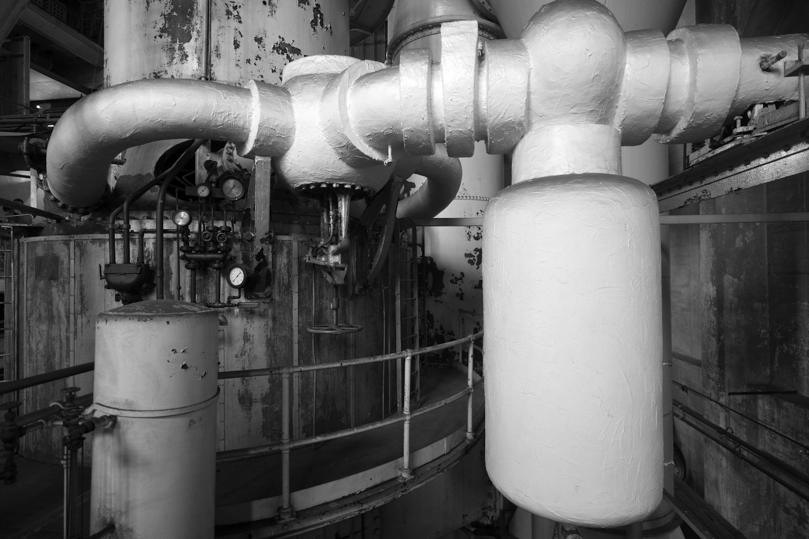 A black and white view inside a plant room — large lagged pipes elbowing overhead into a riser, a rank of pressure gauges and isolation valves at waist height, a handrailed walkway curving around the vessel behind