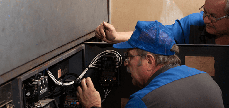 Two service technicians in blue workwear at an opened control cabinet, one holding a bundle of wiring while the other reaches above
