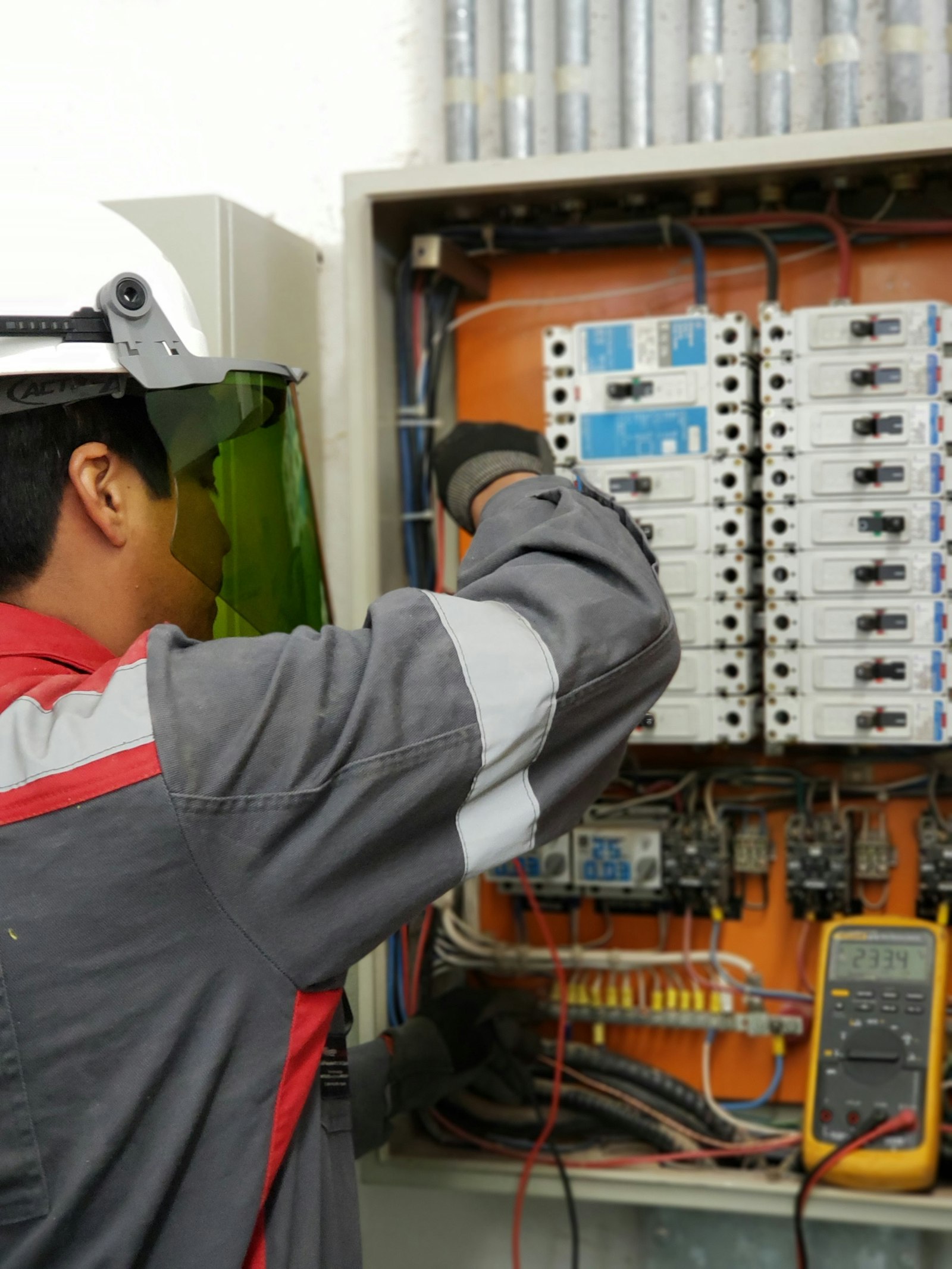 An electrician in a hard hat and green arc-flash face shield reaching into an open control panel, a yellow multimeter reading 233 volts clipped to the door beside rows of breakers and contactors
