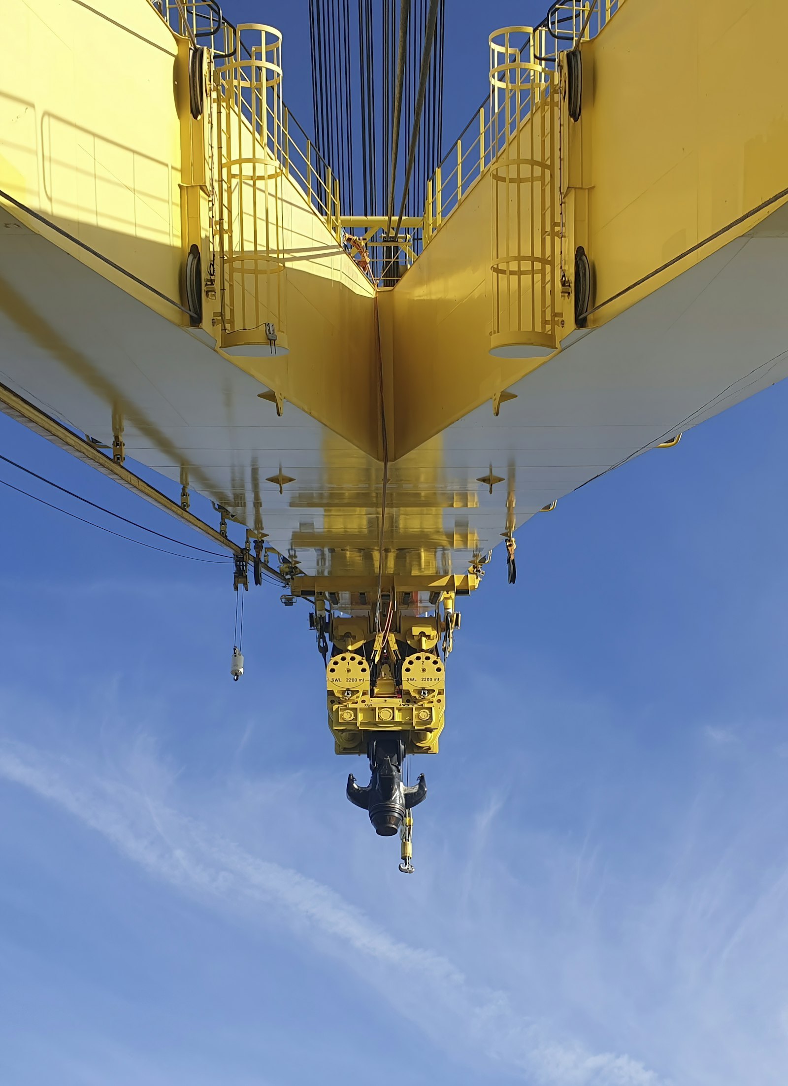 Looking up at the underside of a yellow crane boom against a clear blue sky, its hook block hanging empty on the fall, the rated load stencilled on the sheave housing