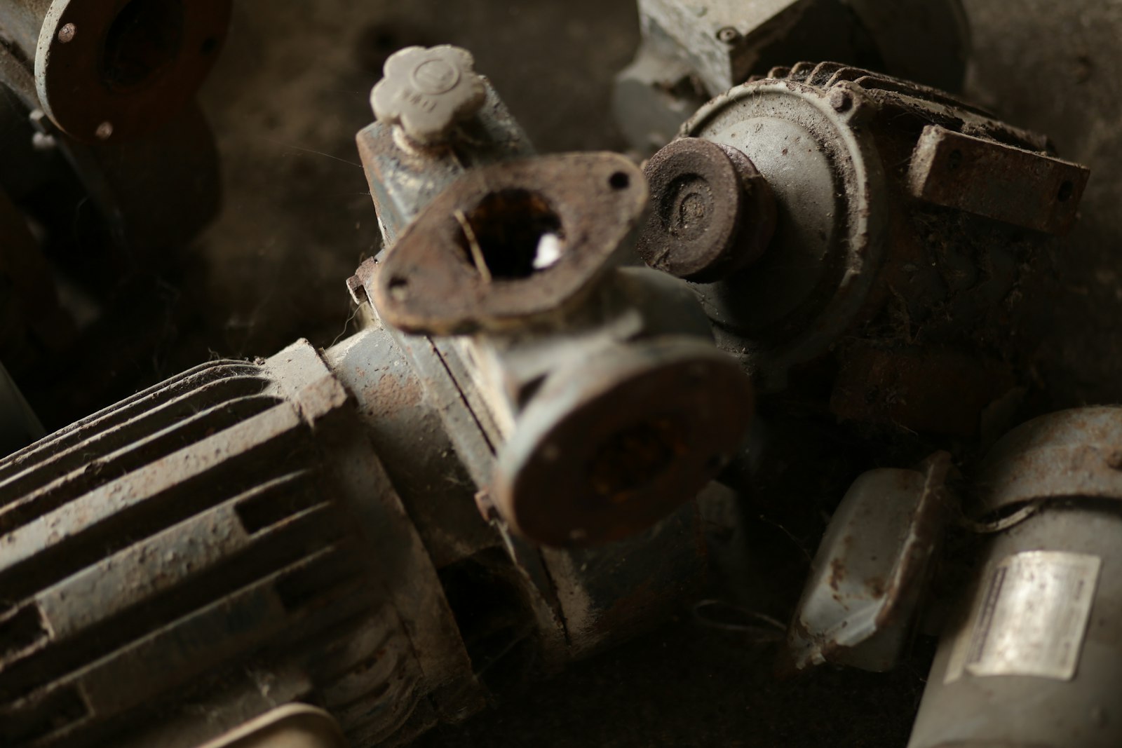 A pile of small electric motors and pump bodies heaped on a concrete floor, thick with dust and cobwebs, flange faces rusted and one coupling missing entirely