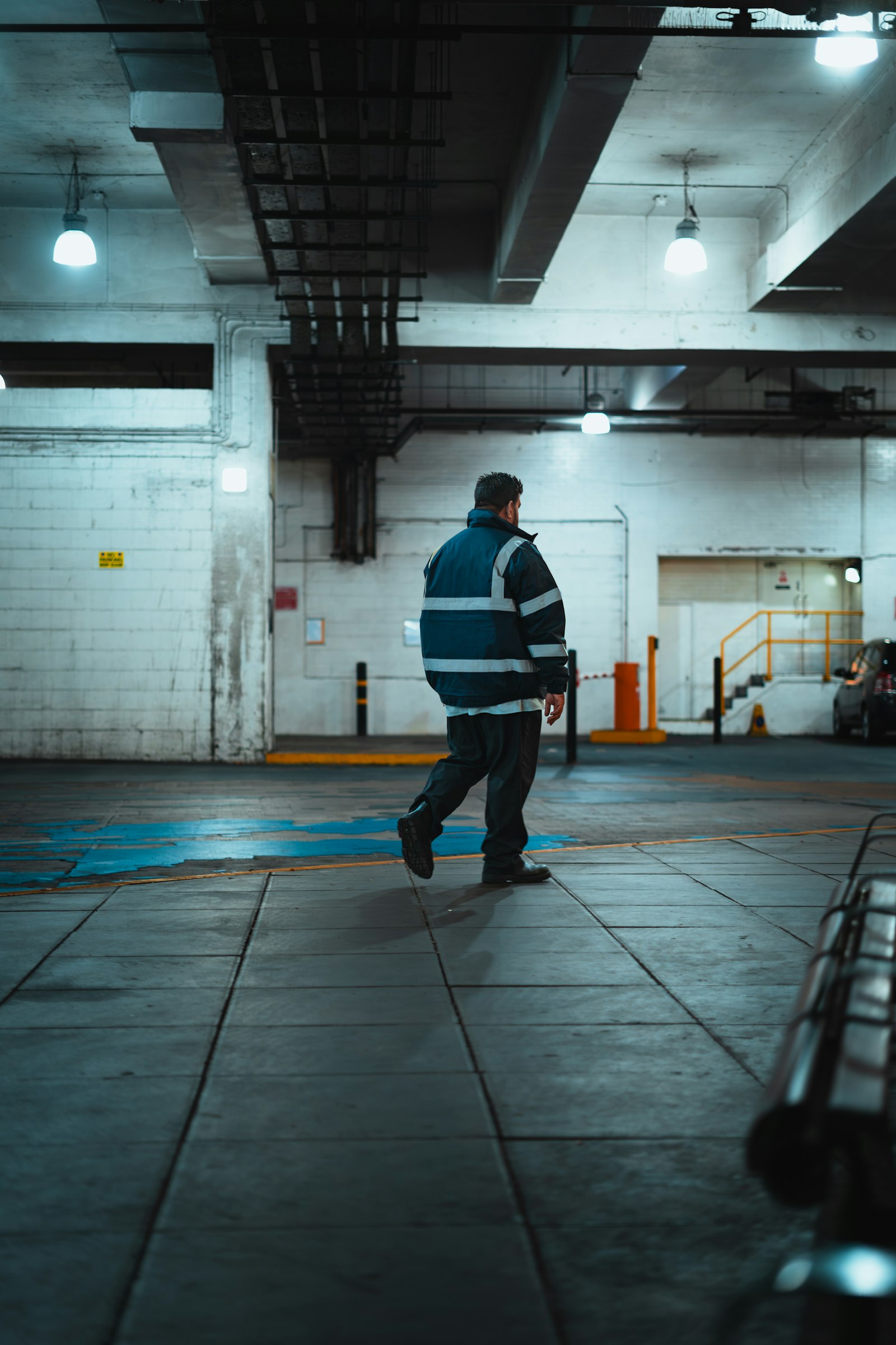 A lone worker in a hi-vis jacket walking across an empty concrete service level at night, lit by widely spaced high-bay fittings, cable tray and pipework running overhead