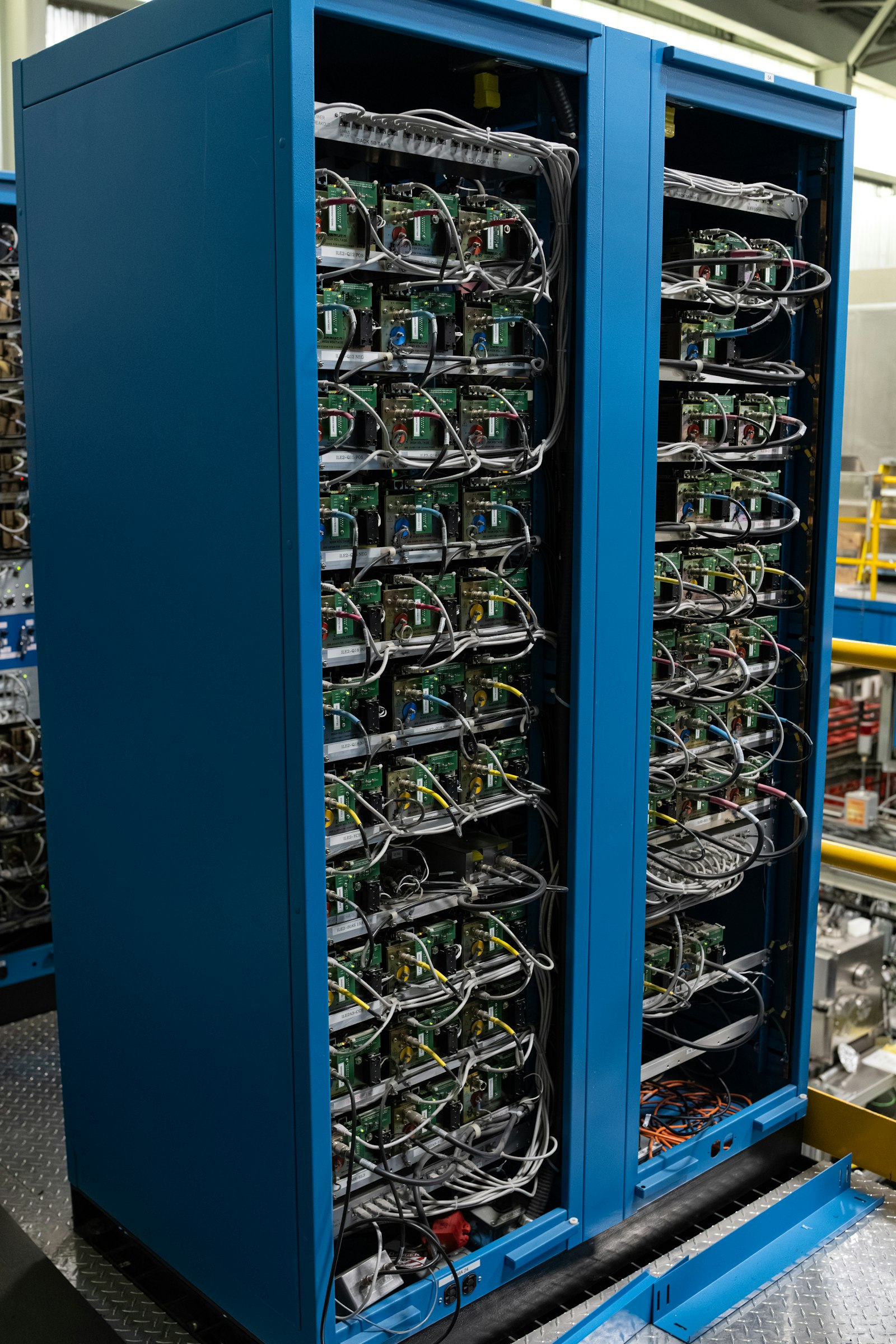 Two blue equipment cabinets standing open on a plated floor, every shelf packed with green circuit cards and a dense harness of grey and coloured cabling