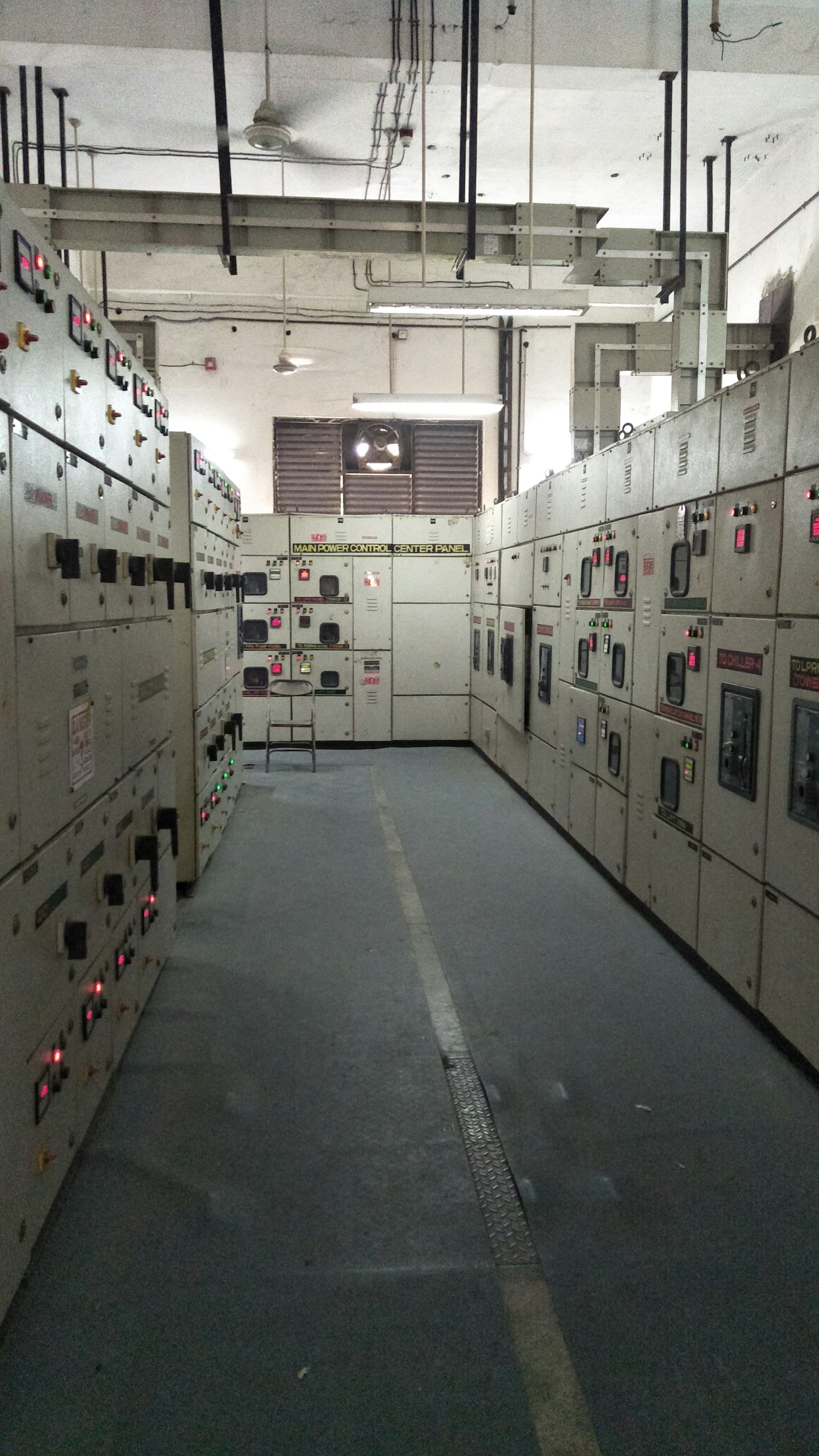 A long aisle between two facing rows of low-voltage switchgear cubicles, indicator lamps lit red and green down both sides, a section at the far end labelled Main Power Control Center Panel