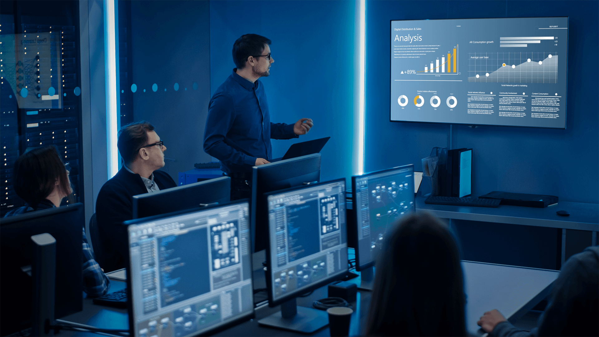 A team in a dark control room watching a colleague present an analysis dashboard on a wall screen, code and charts on the monitors in front of them