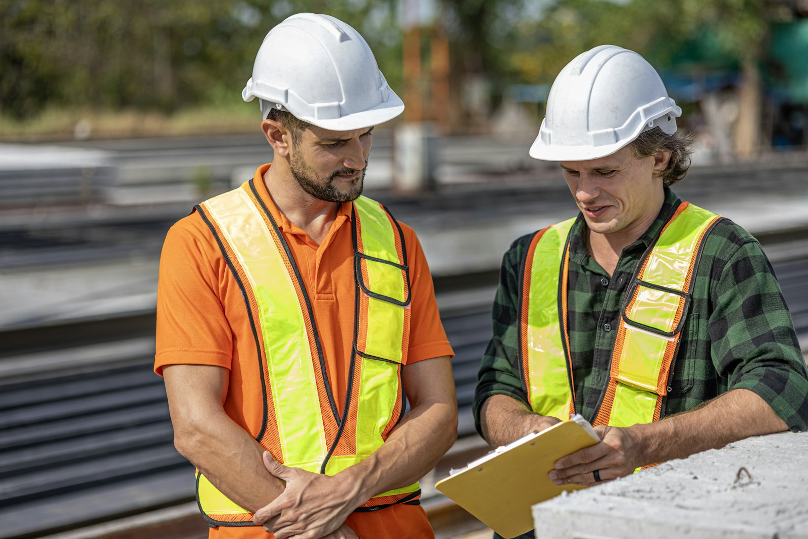 Two workers in hi-vis vests and white hard hats standing together over a yellow clipboard on a job site, one writing while the other reads
