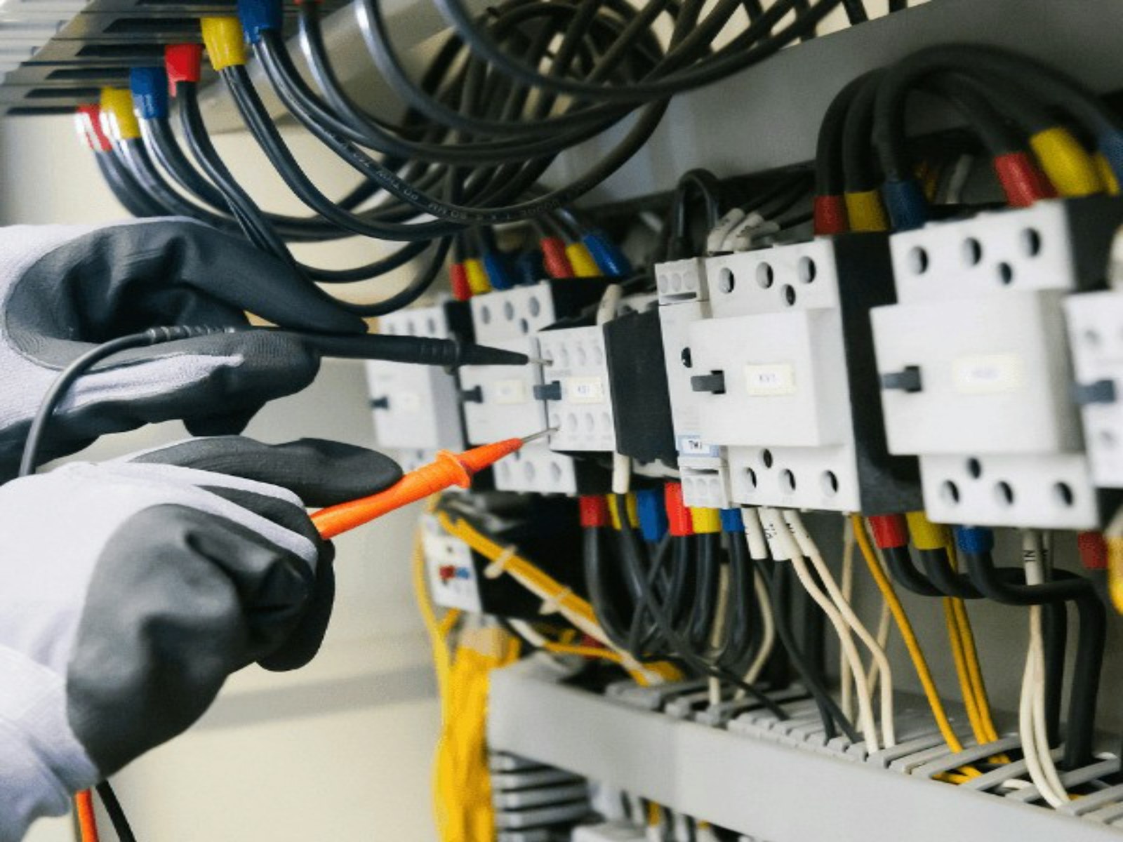 Two gloved hands holding meter probes against a DIN rail of white contactors and relays, the panel behind them full of looped black, red and yellow control wiring
